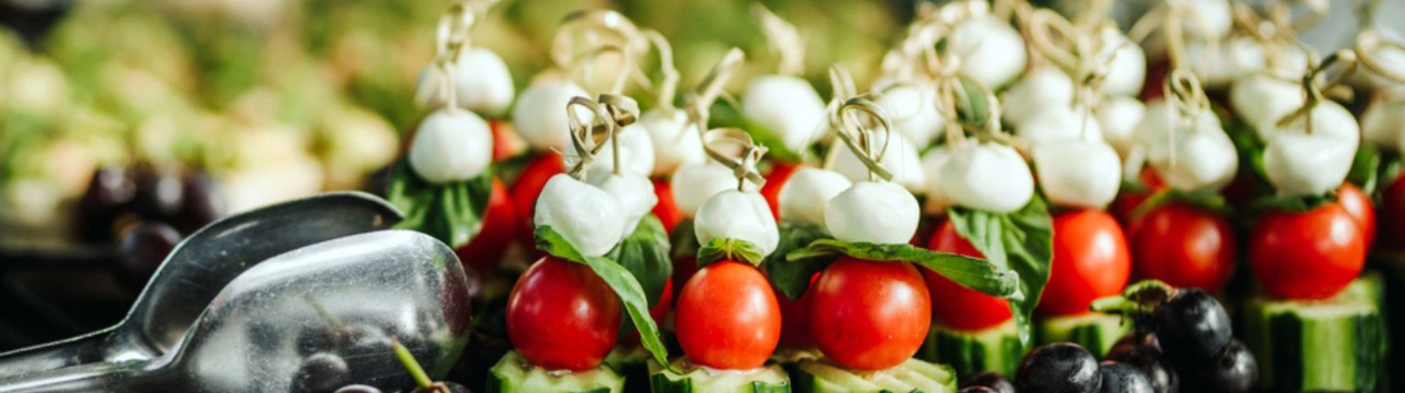 Brochettes de tomates cerises, billes de mozzarella et feuilles de basilic disposées sur un plateau avec des olives et des cuillères métalliques.