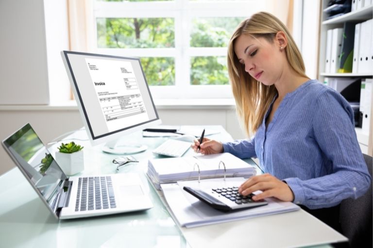 Une femme faisant de la comptabilité avec une calculatrice et deux écrans d'ordinateur