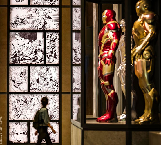 Photographie de statue de trois différentes combinaisons du super-héro Iron-Man de l'univers cinématographique Marvel®, et mur habillé d'extraits de bandes-déssinées
